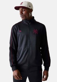 Giacca nera con zip e colletto alto, dettagli rossi e logo ricamato. Abbinate a un cappellino bianco e bordeaux con visiera curva.