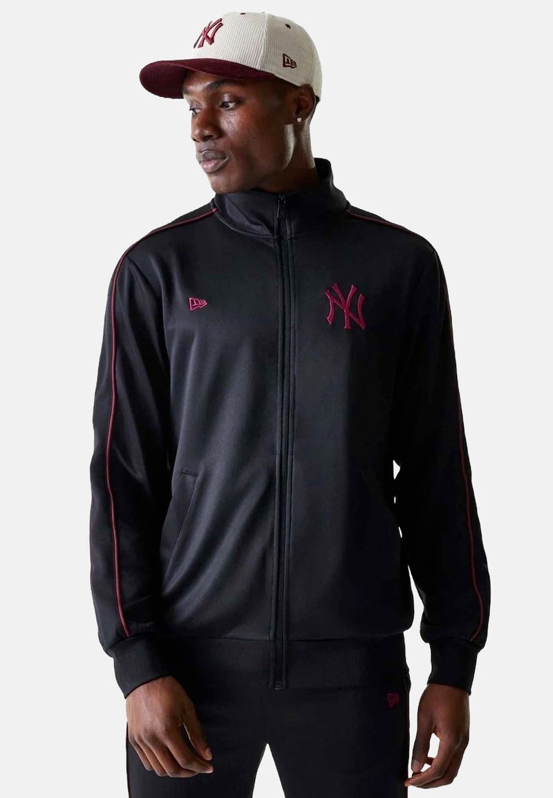 Giacca nera con zip e colletto alto, dettagli rossi e logo ricamato. Abbinate a un cappellino bianco e bordeaux con visiera curva.