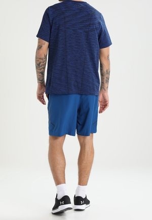 Un homme portant un t-shirt bleu marine, un short bleu, des chaussettes blanches et des baskets noires Under Armour, debout, de dos face à la caméra.