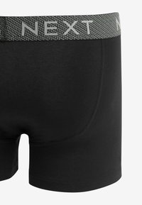 Schwarze Boxer Shorts mit einem strukturierten grauen Bund, auf dem das Wort "NEXT" steht. Hergestellt aus glattem, dehnbarem Material mit einer taillierten Passform.