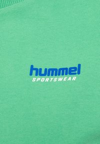 Niebieskie logo tekstowe "hummel" i białe "SPORTSWEAR" na zielonym, teksturowanym tle materiału.