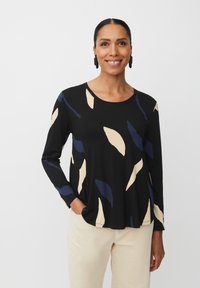 Zwarte top met lange mouwen met beige en marineblauwe bladerenpatronen. Ronde halslijn, zacht materiaal en een ontspannen pasvorm, gecombineerd met lichtgekleurde broeken.