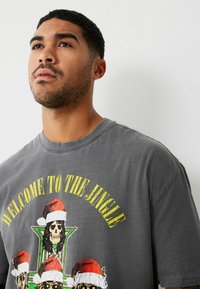 Szürke, túlméretezett pamut póló, amelyen koponyákat ábrázoló grafika látható, karácsonyi sapkában, élénk sárga "WELCOME TO THE JUNGLE" felirattal.