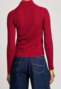 Roter gerippter Strickpullover mit hohem Kragen, langen Ärmeln und tailliertem Bund, kombiniert mit dunklen Denim-Jeans mit Gesäßtaschen.
