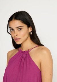 Vestido fucsia de cuello halter con escote fruncido, confeccionado en tejido ligero, con tirantes finos y un ajuste relajado. Pendientes de aro dorados.