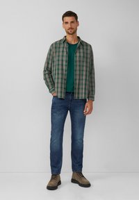 Karierte Button-up-Hemd in Grün, Beige und Braun, über einem türkisfarbenen T-Shirt getragen. Kombiniert mit schmal geschnittenen blauen Jeans und beigen Schnürstiefeln.