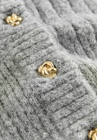 Grå strikket beklædning med et tekstureret mønster og guldfarvede hardwaredetaljer. Nærbillede viser blødt, fuzzy garn og dekorative knapper.