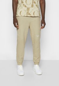 Beiga bomullssweatpants med elastiska muddar, prydda med en vit logotyp på vänster lår och bärs med vita sneakers och en mönstrad skjorta.