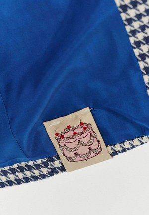 Modrá látka s okrajem v kostkované houndstooth vzoru. Štítek zobrazuje ilustraci růžového dortu s červenými třešněmi navrchu a krémovými detaily.