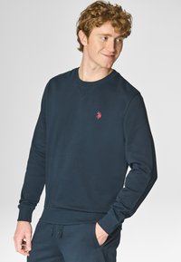 Felpa navy con collo rotondo e polsini a costine. Presenta un piccolo logo rosso sul lato sinistro del petto. Tessuto morbido, vestibilità comoda.