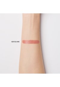 Échantillon de produit à lèvres étiqueté "829 BLAME" sur un bras à la peau claire, montrant une texture lisse et mate dans une couleur rose-brun atténuée.