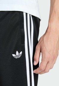 Black athletic pants with white side stripes and a small white Adidas logo. Smooth fabric texture, accentuated by a hand touching the material.