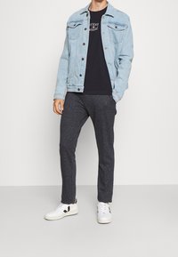 Giubbotto in denim blu chiaro con dettagli in bottone, t-shirt nera sotto, pantaloni della tuta grigi scuri e sneakers bianche con accenti neri.