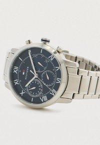 Ezüst fém karóra fekete számlappal, római számokkal jelzett óranézők, három al-számlappal, amelyek a napot, a dátumot és a 24 órás időt mutatják, valamint láncos szíjjal.