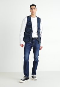 Denim vest with multiple pockets, layered over a white long-sleeve shirt, paired with blue jeans and black canvas sneakers.