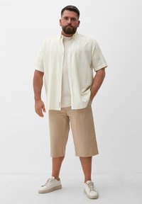 Korte mouwen shirt in lichtgeel met witte strepen, gecombineerd met beige shorts tot de knie en witte sneakers. Soepele stof, met een knoopsluiting aan de voorkant.