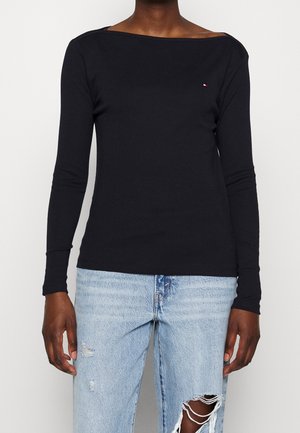 Personne portant un haut noir ajusté à manches longues et un jean bleu clair déchiré, debout devant un fond uni.