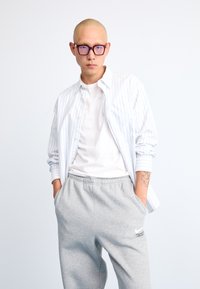 Chemise blanche à rayures bleues sur un t-shirt blanc, associée à un pantalon de survêtement gris arborant un logo. Lunettes avec une monture violette.