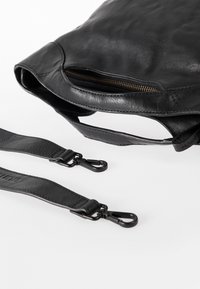 Sac en cuir noir avec un dessus zippé, texture lisse, bords arrondis et sangles détachables avec clips métalliques pour une fixation sécurisée.