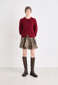 Cardigan en laine rouge avec encolure ronde et fermeture à boutons, associé à une robe évasée à imprimé léopard et des bottes montantes marron.