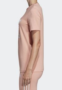 Person som bär en ljusrosa Adidas t-shirt och matchande leggings, stående på sidan mot en enfärgad bakgrund.