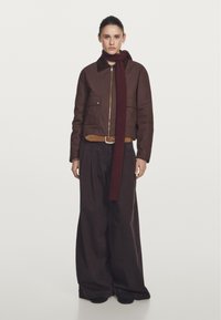 Brązowa krótka kurtka z zamkiem z przodu i dwiema kieszeniami, w połączeniu z szerokimi czarnymi spodniami. Ciemnoczerwona prążkowana szalik owinięty wokół szyi.