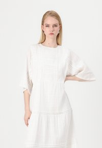 Bela bombažna obleka s kratkimi, širokimi rokavi, teksturiranim vzorcem, scallop robom in zapletenimi šivi ob bodice.
