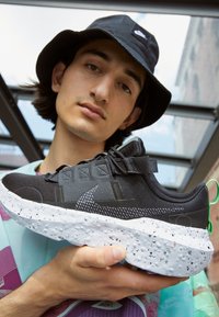 Young man wearing a black Nike bucket hat holds a black and white Nike sneaker with speckled sole, shown close to the camera.