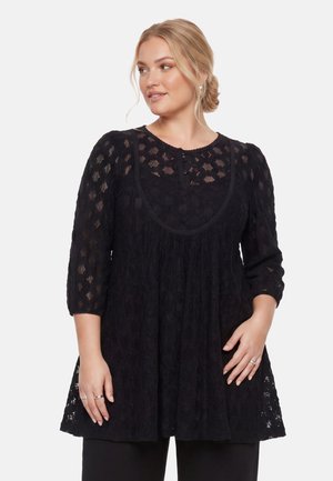 Femme aux cheveux blonds en chignon portant un haut en dentelle noire ample avec des manches trois-quarts, regardant sur le côté avec les mains reposant sur la taille.