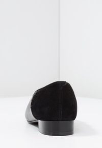 Svart sko med en mockautsida och en slät läderdetalj. Låg, rund klack och en elegant, minimalistisk design.