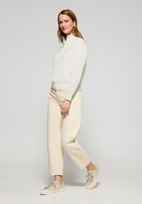 Licht beige sweatshirt met halve rits en geribde mouwen, gecombineerd met off-white wijde jeans en lichte sneakers, geplaatst tegen een neutrale achtergrond.