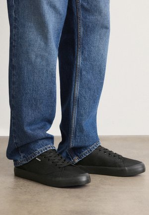 Ténis pretos de lona com solas de borracha e bico redondo. Design com atacadores e pequeno detalhe em branco do logótipo na lateral. Usados com jeans de ganga azuis.