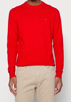 Hombre con suéter rojo brillante de manga larga con pequeño logo en el pecho y pantalones de pana beige claro, de pie frente a fondo blanco.