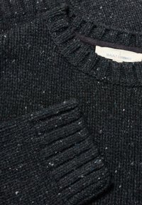 Tamno sivi pleteni pulover s rebrastim izrezom i manžetama. Ima bijele i tirkizne flekice kroz cijelo platno. Na etiketi piše "Seasalt Cornwall."