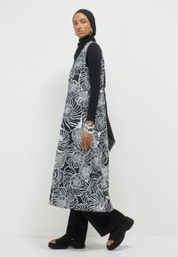 Donna che indossa un turbante nero, un top nero a maniche lunghe, pantaloni neri, bracciali d'argento e un abito senza maniche con motivo floreale in bianco e nero.