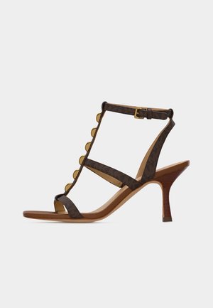 Hnedá kožená sandála na vysokom opätku so zlatými gombíkmi, členkovým remienkom a dreveným ihličkovým opätkom na bielom pozadí.