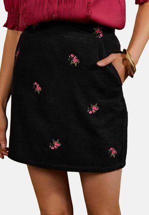 Sort kordnederdel med små lyserøde blomsterbroderier, båret med en rød bluse, hånd i lommen og gyldne armbånd på håndleddet.