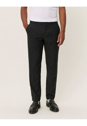 Homme portant un pantalon de costume gris foncé, une chemise blanche et des chaussures en cuir noires, debout les mains dans les poches sur un fond uni.