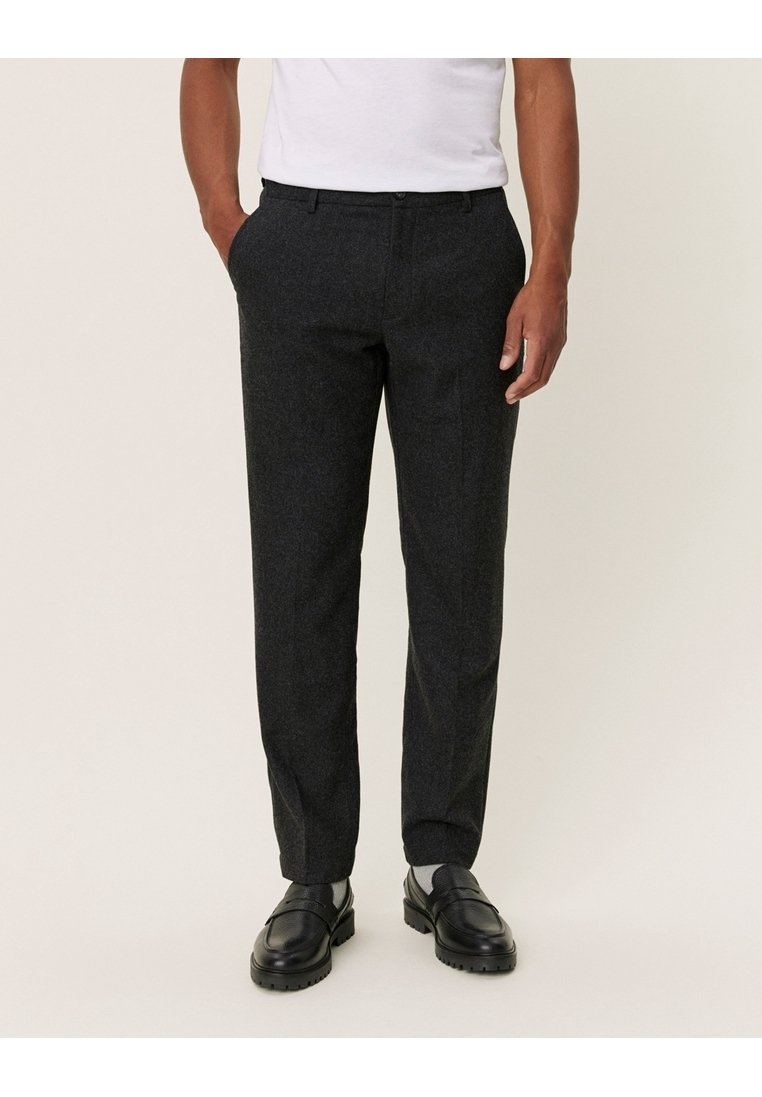 Homme portant un pantalon de costume gris foncé, une chemise blanche et des chaussures en cuir noires, debout les mains dans les poches sur un fond uni.
