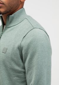Lichtgroene zip-up sweater met een ribbelkraag en textuurstof. Bevat een subtiele logopatch op de borst en een getailleerde silhouette.