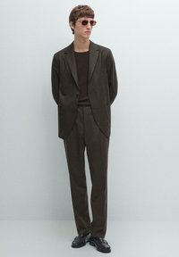 Man wearing dark brown suit with matching sweater and black leather loafers, standing against plain light gray background.