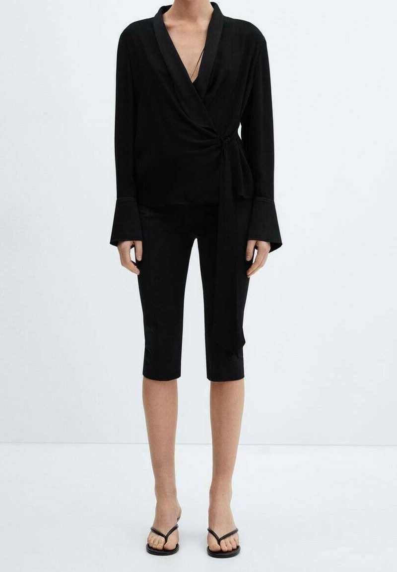 Personne portant un chemisier noir à manches longues de style enveloppant, un short noir longueur genou, et des sandales plates noires à bouts ouverts sur fond uni.