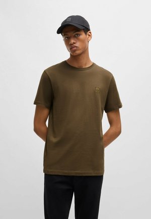 Olivengrünes Baumwoll-T-Shirt mit Rundhalsausschnitt und kurzen Ärmeln, versehen mit einem kleinen quadratischen Logo-Patch auf der Brust, kombiniert mit einer schwarzen Kappe.
