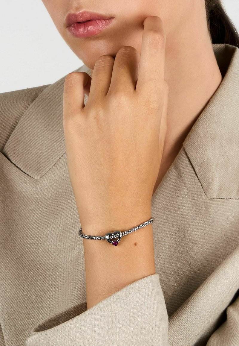 Bracciale in argento con un charm a forma di cuore, decorato con un accento in pietra preziosa rossa, attaccato a una catena intrecciata. Sfondo in tessuto texturizzato.