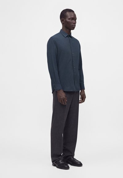 Homme debout de profil portant une chemise bleu foncé boutonnée, un pantalon large foncé et des chaussures en cuir noires sur un fond blanc.