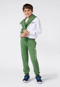 Zēns, tērpies baltā sporta krekls un zaļos sporta biksēs, ar atbilstošu zaļu džemperi, kas uzlikts uz pleciem, stāv atviegloti, rokas saliktas kopā.