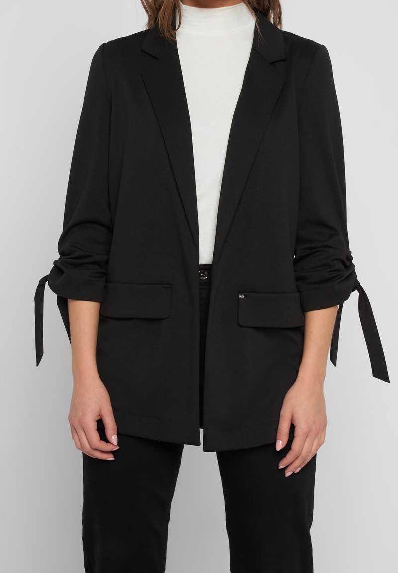 Blazer noir en tissu doux, avec un col cranté, une ouverture avant sans fermeture, des manches retroussées avec liens, et deux poches avant.