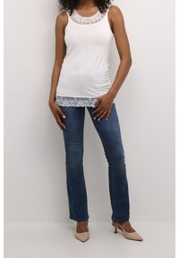 Vit tanktopp med spetskant vid halslinning och nederkant, kombinerad med mörkblå utsvängda jeans och neutrala klackar.