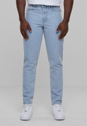 Mann iført lyseblå slim-fit jeans og hvite sneakers, står med hendene avslappet ved siden av seg mot en ensfarget grå bakgrunn.
