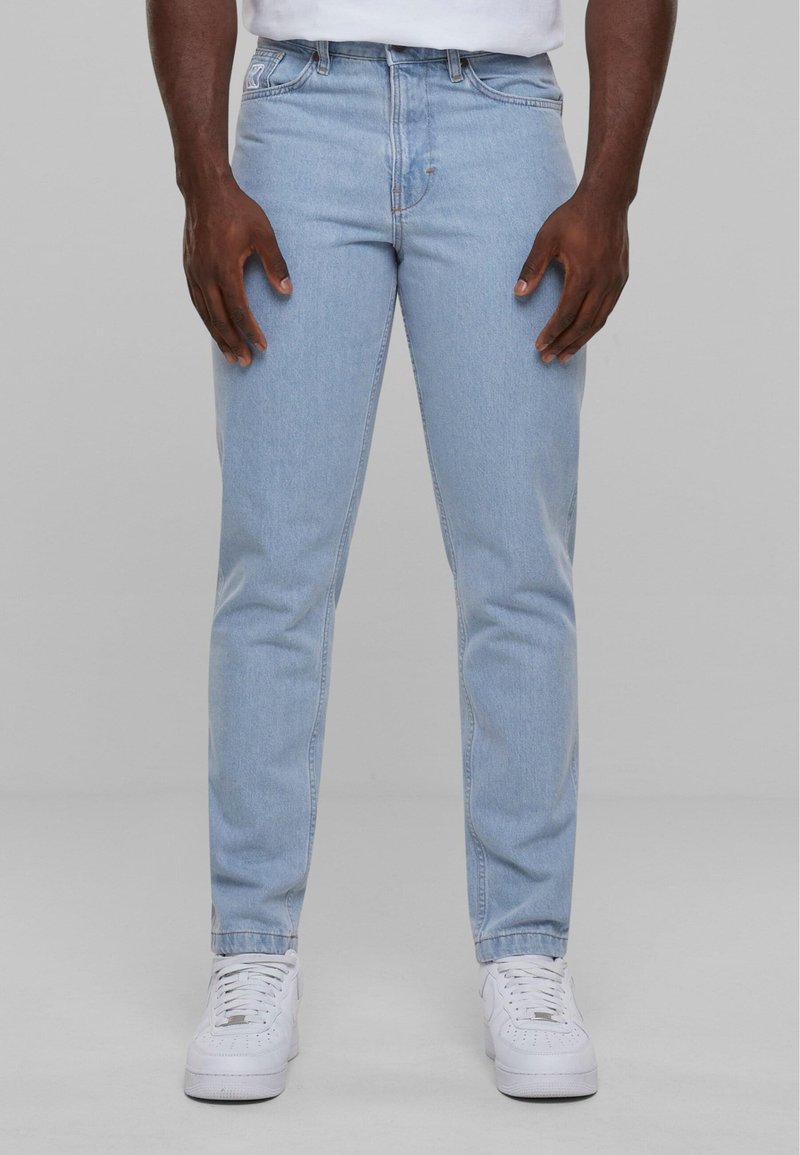 Uomo in jeans slim-fit azzurri chiari e sneakers bianche, in piedi con le mani rilassate ai lati davanti a uno sfondo grigio uniforme.